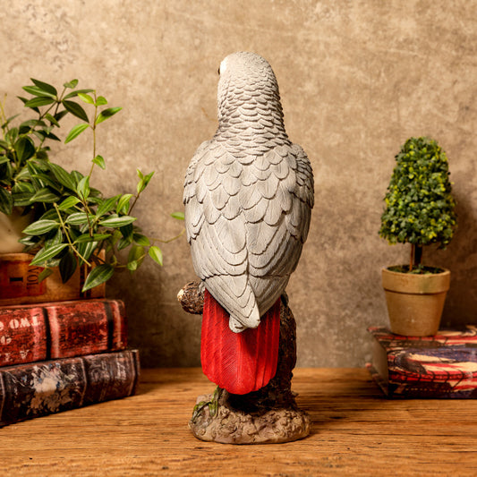 Statue of a parrot with red tail on a wooden surface with plants and books in the background