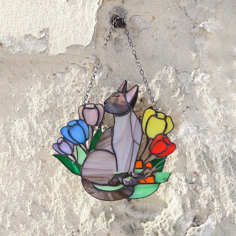 Stained glass suncatcher of a cat and flowers hanging on a textured wall.
