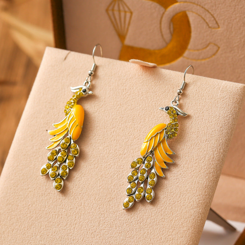Pair of peacock feather earrings with yellow and green stones on a beige surface.