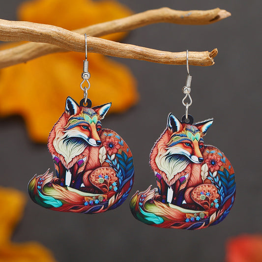 Colorful fox earrings hanging from a branch with a blurred autumn leaf background.