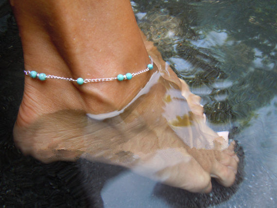 Close-up of a foot with a turquoise beaded anklet standing in water.