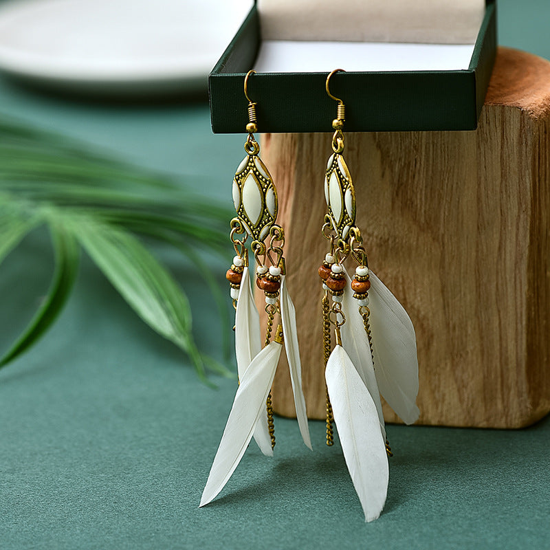 Feather earrings with gold accents on a green surface with a wooden block and plant leaves in the background from Nested Wisdom