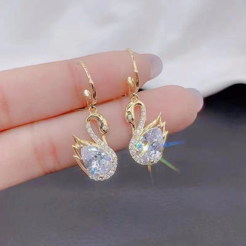 Gold Swan-shaped earrings held between fingers against a neutral background