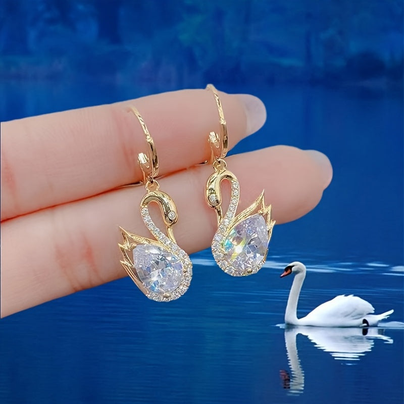 Gold swan earrings held by a hand with a swan in the background on water.