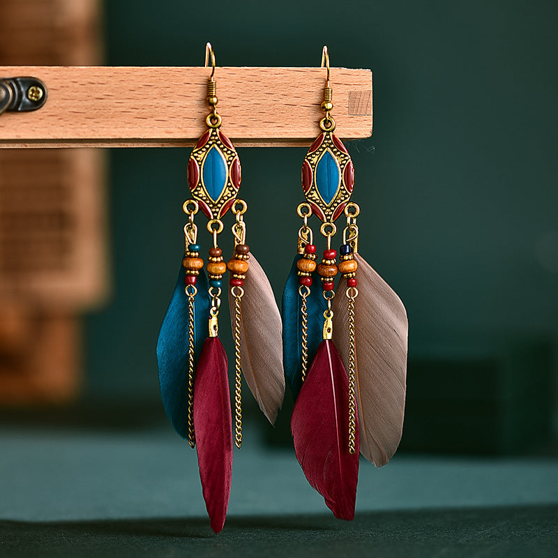 Feather earrings with blue, red, and gold details on a wooden stand against a dark green background. From Nested Wisdom