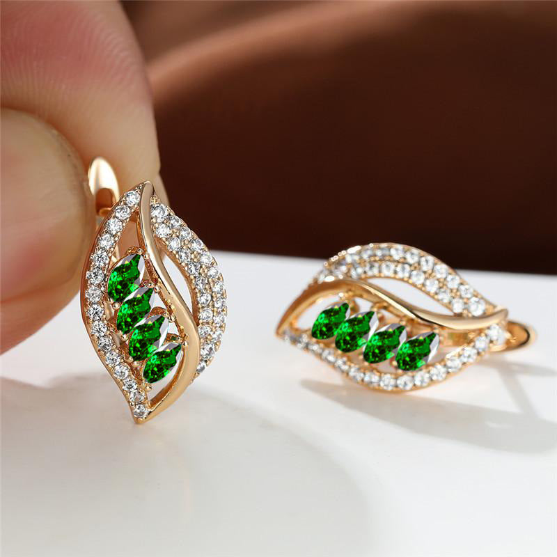 Gold earrings with green gemstones held by a hand on a white surface.