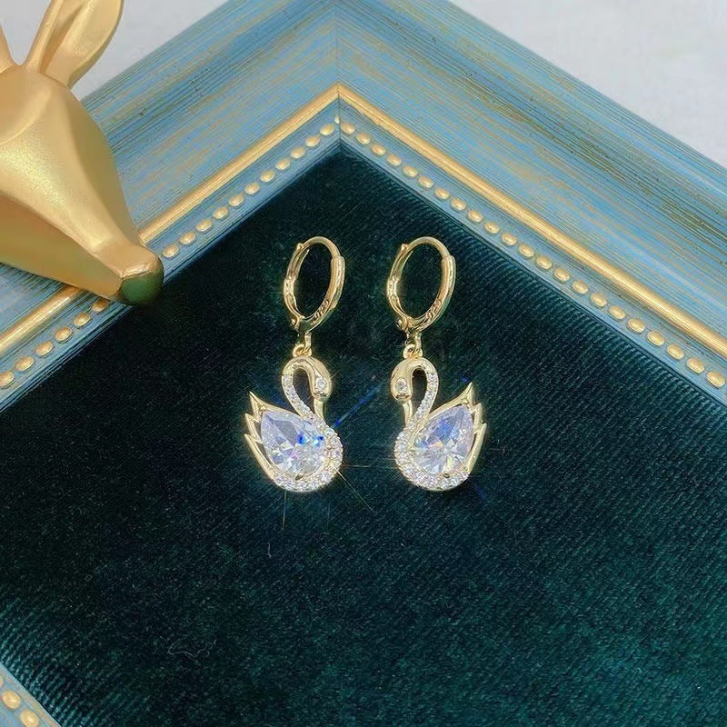 Gold swan-shaped earrings on a green velvet surface with a gold frame.