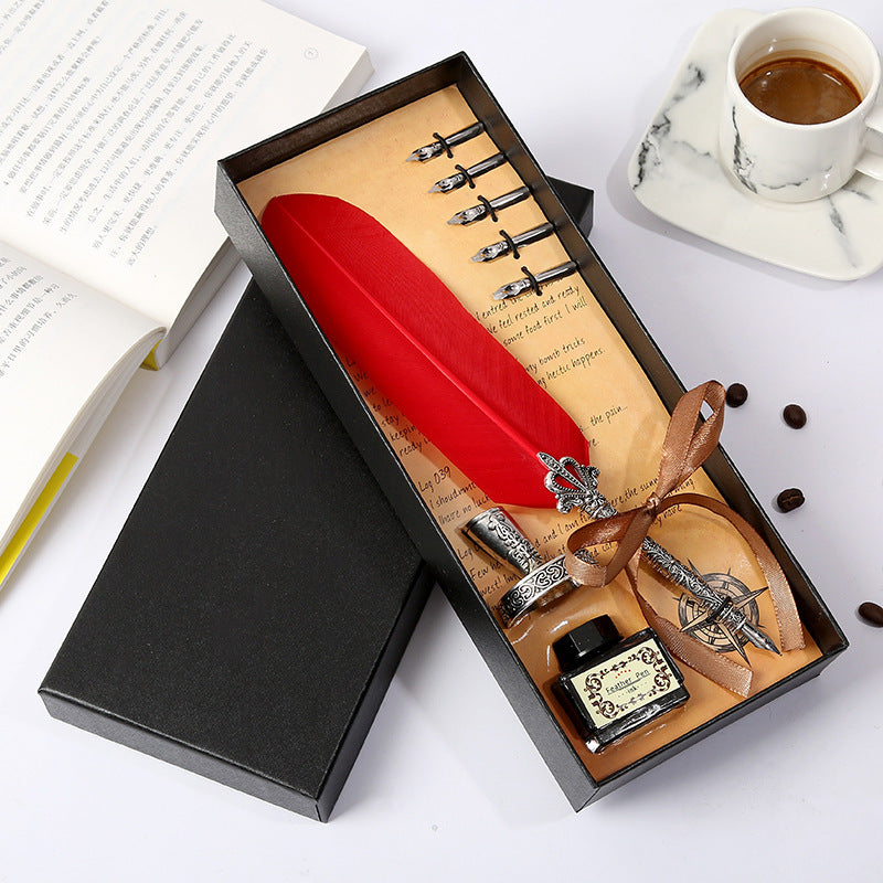 Red feather pen set with ink bottle and nibs in a black box on a white surface.