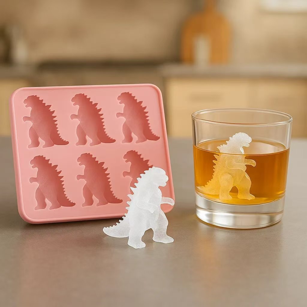 Silicone mold shaped like Godzilla next to a glass of orange liquid on a wooden surface.