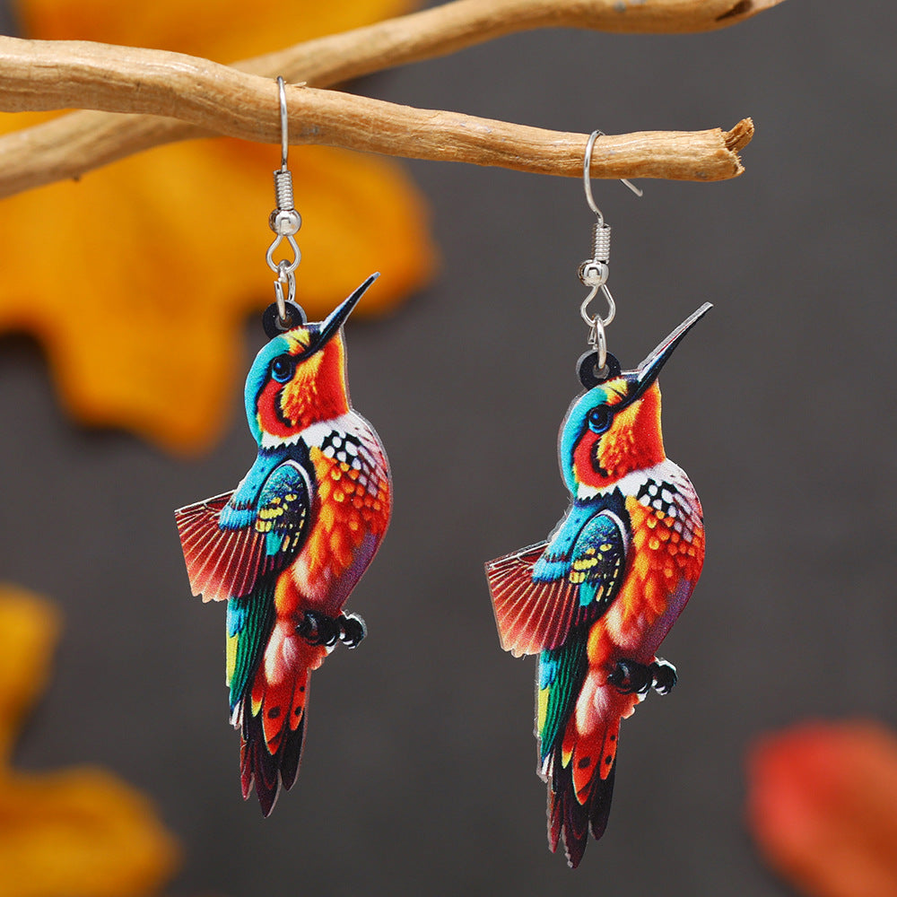 Colorful hummingbird-shaped earrings hanging on a branch with a blurred floral background.