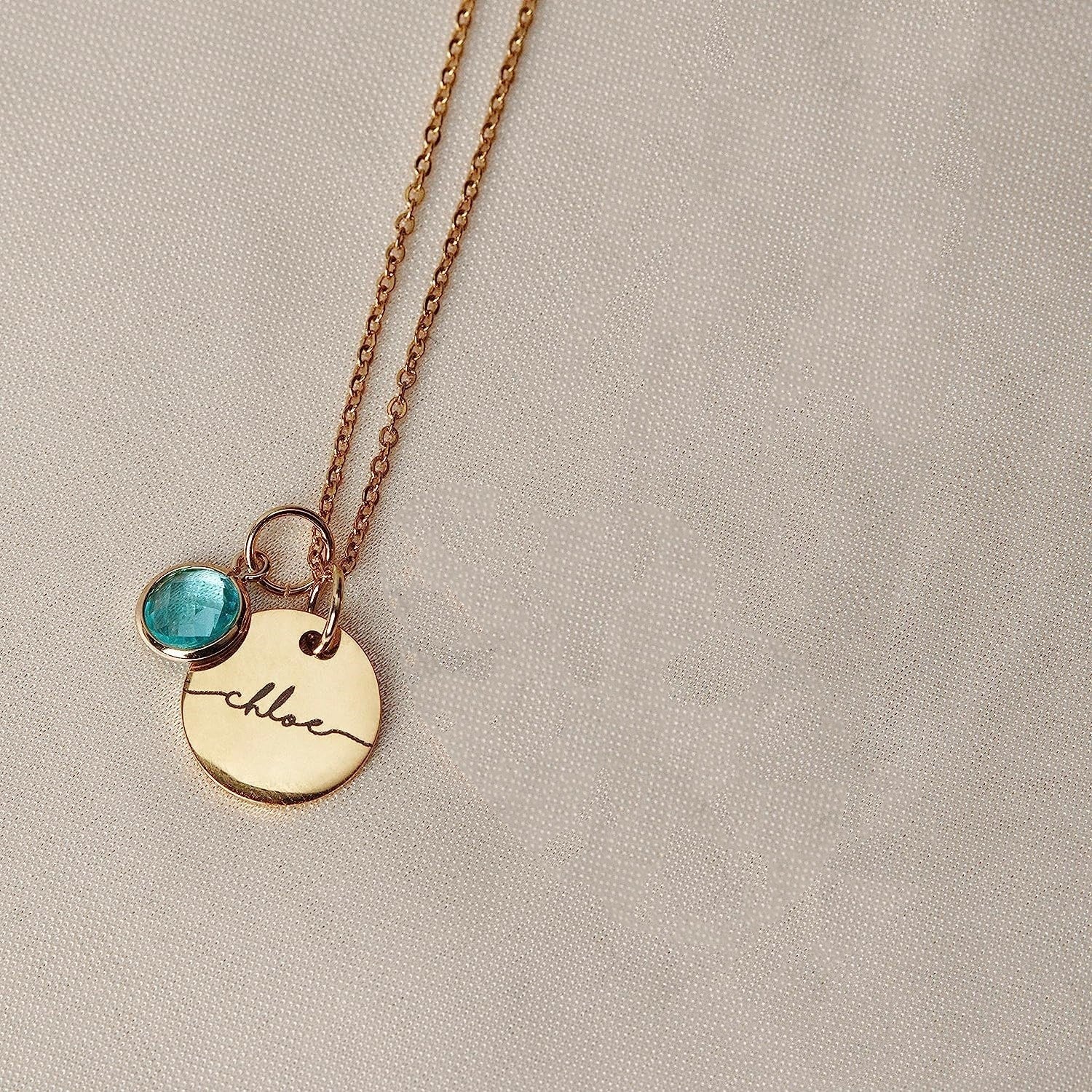 Gold necklace with a blue gemstone and engraved name on a beige background