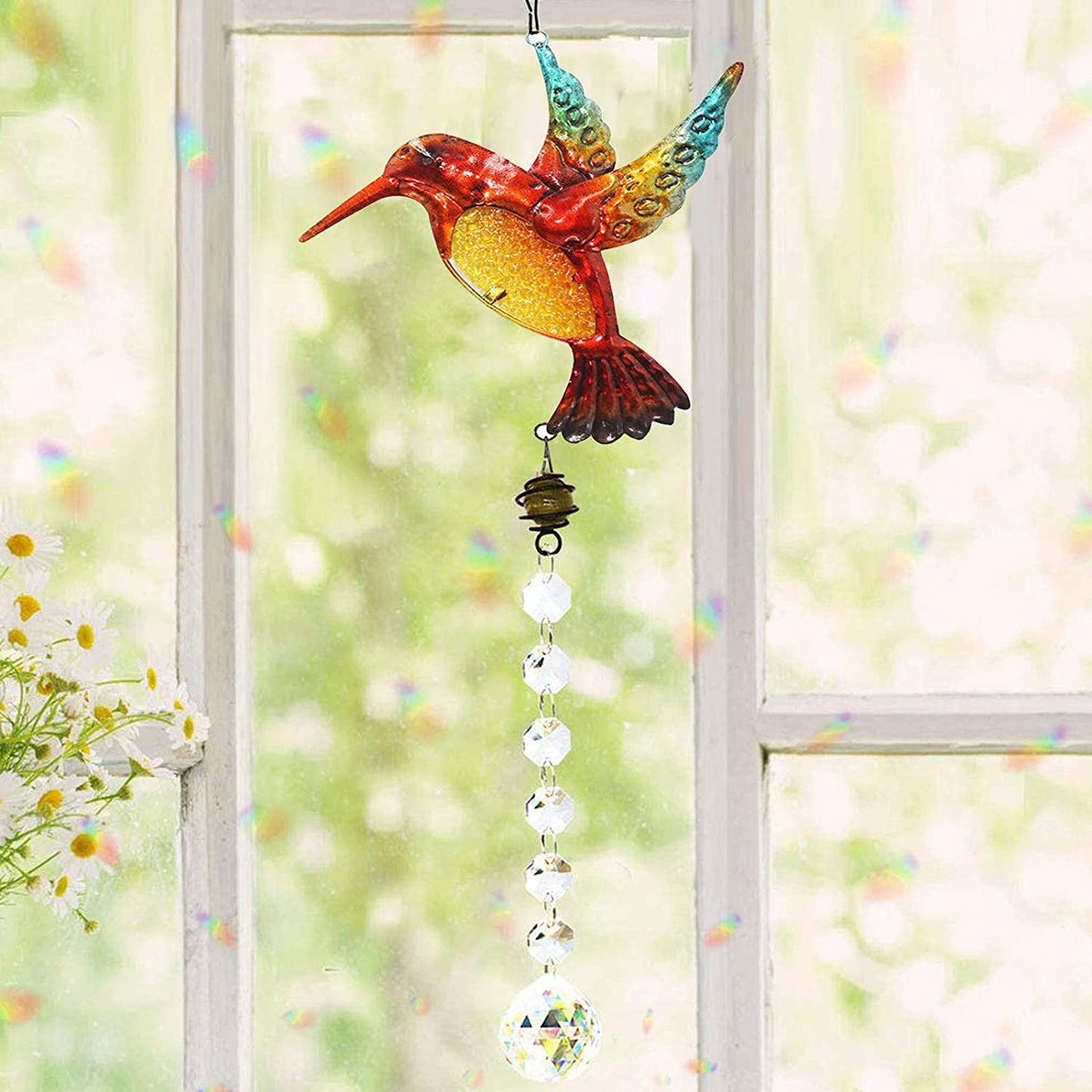 Colorful hummingbird-shaped glass ornament hanging by a window with a blurred garden background.