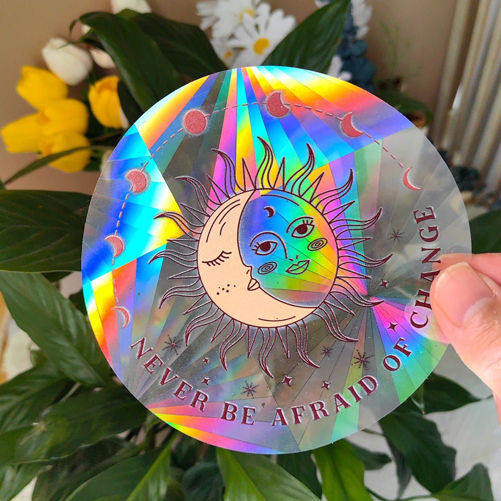 Holographic sun and moon sticker with 'Never be afraid of change' text held in front of greenery.