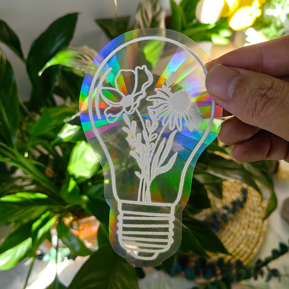 Holographic light bulb-shaped cutout with floral design held by a hand against a plant background