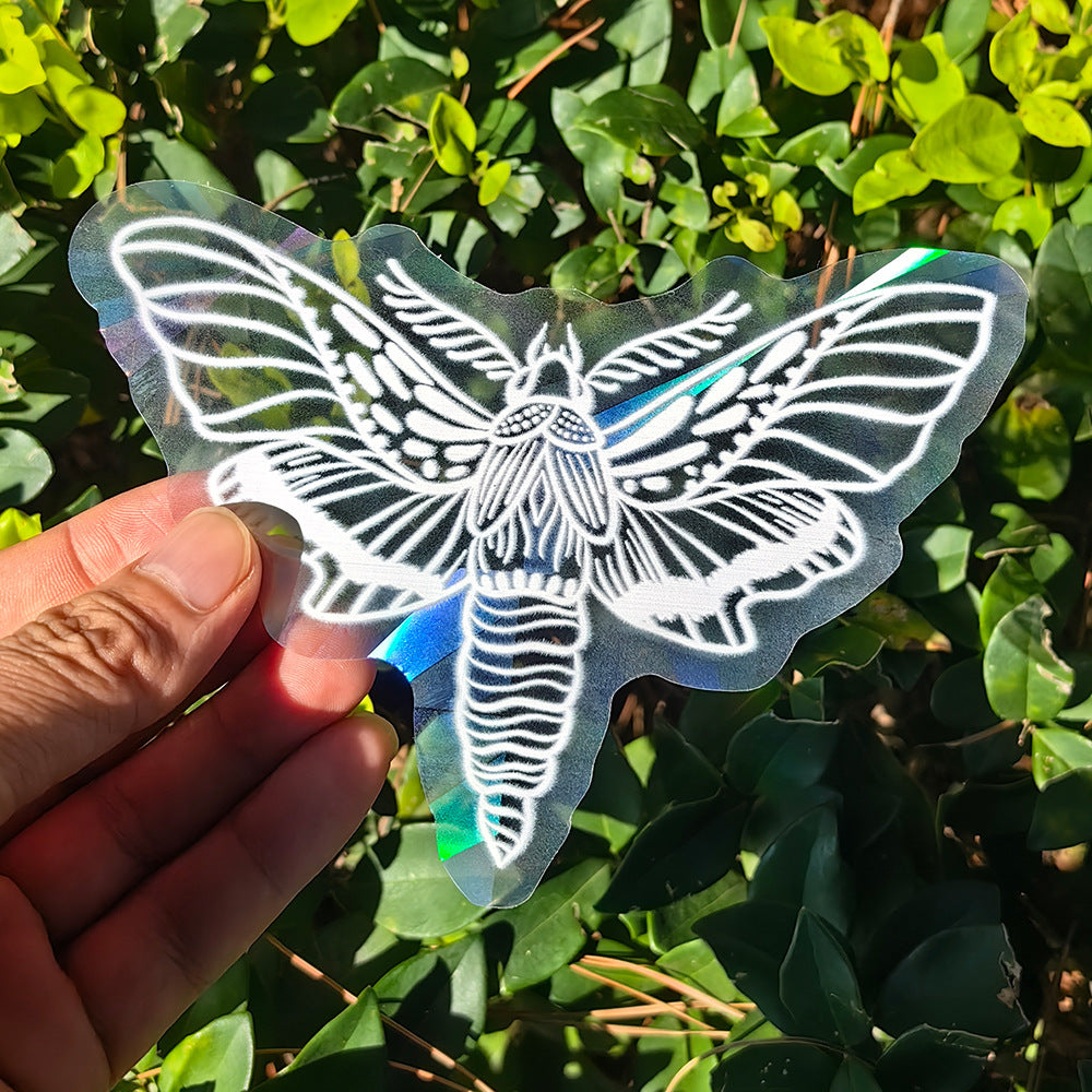 Hand holding a moth-shaped holographic sticker against a green leafy background