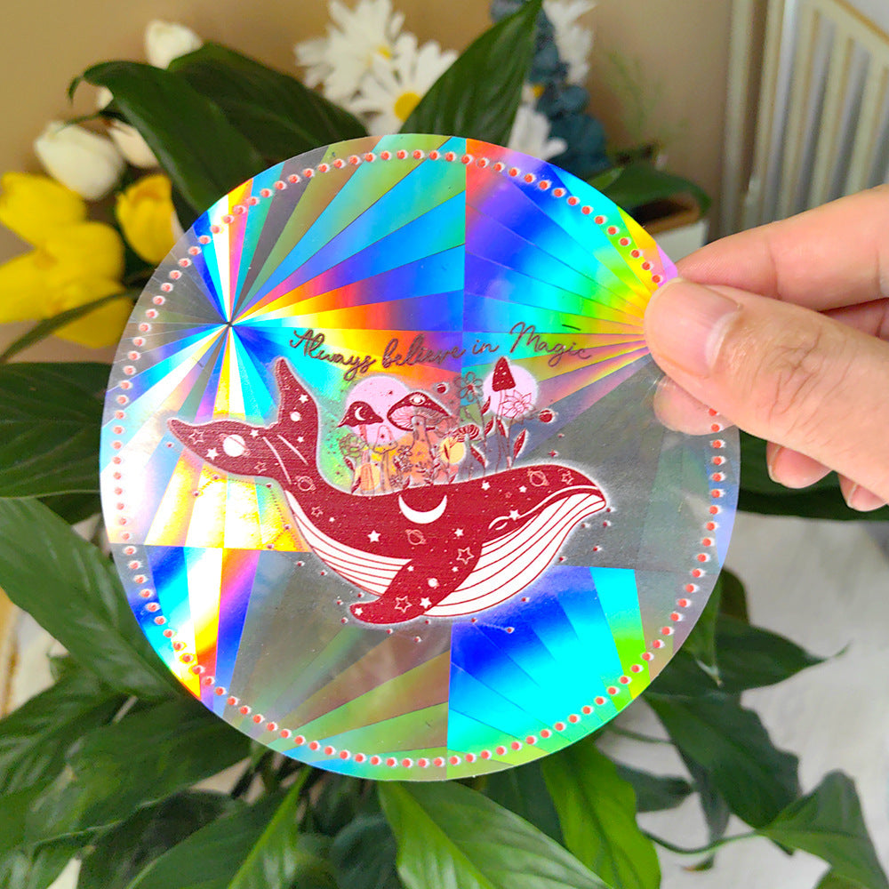Holographic sticker with a whale and fairy design held by a hand, with plants in the background.