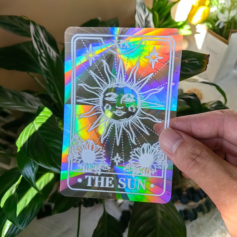 Holographic card with 'The Sun' design held by a hand in front of green plants.