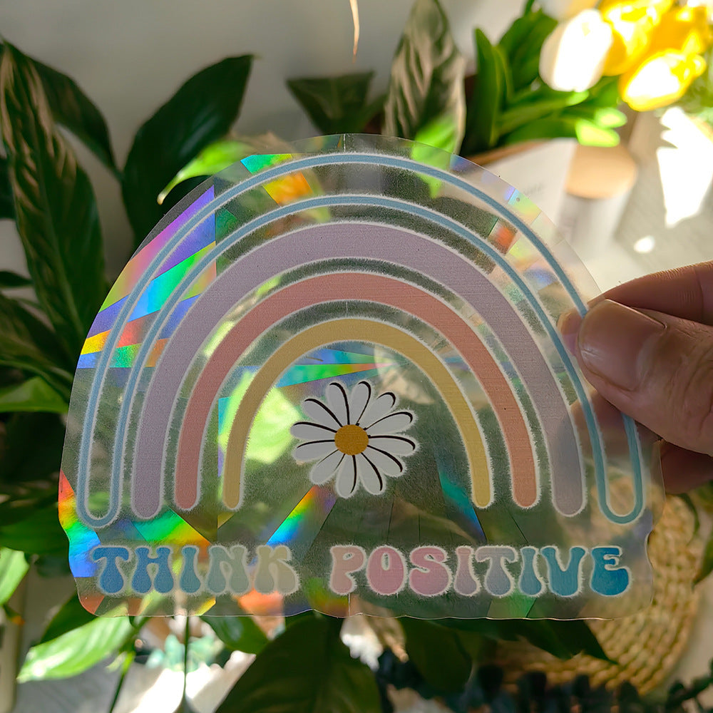 Holographic rainbow-shaped object with a daisy and 'Think Positive' text, held in front of plants.