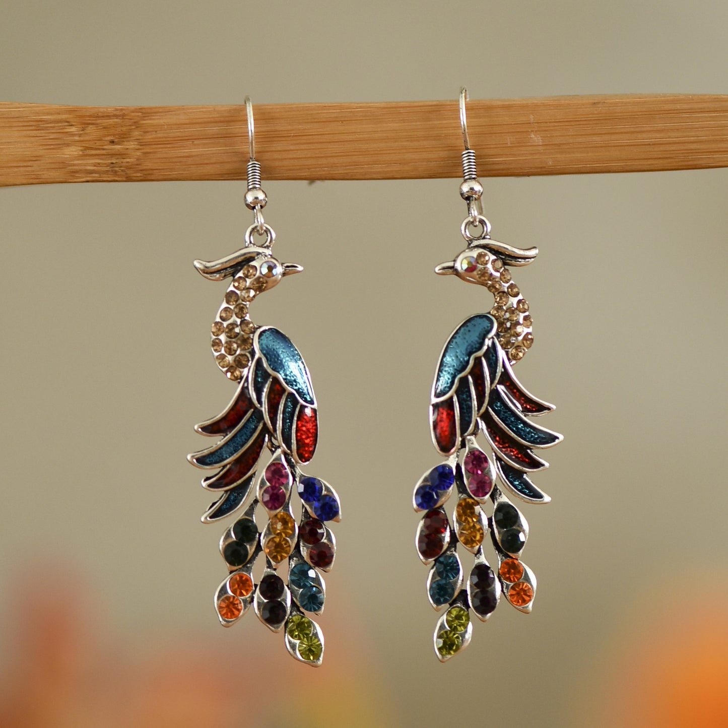 Colorful peacock-shaped earrings hanging on a wooden rod with a blurred background