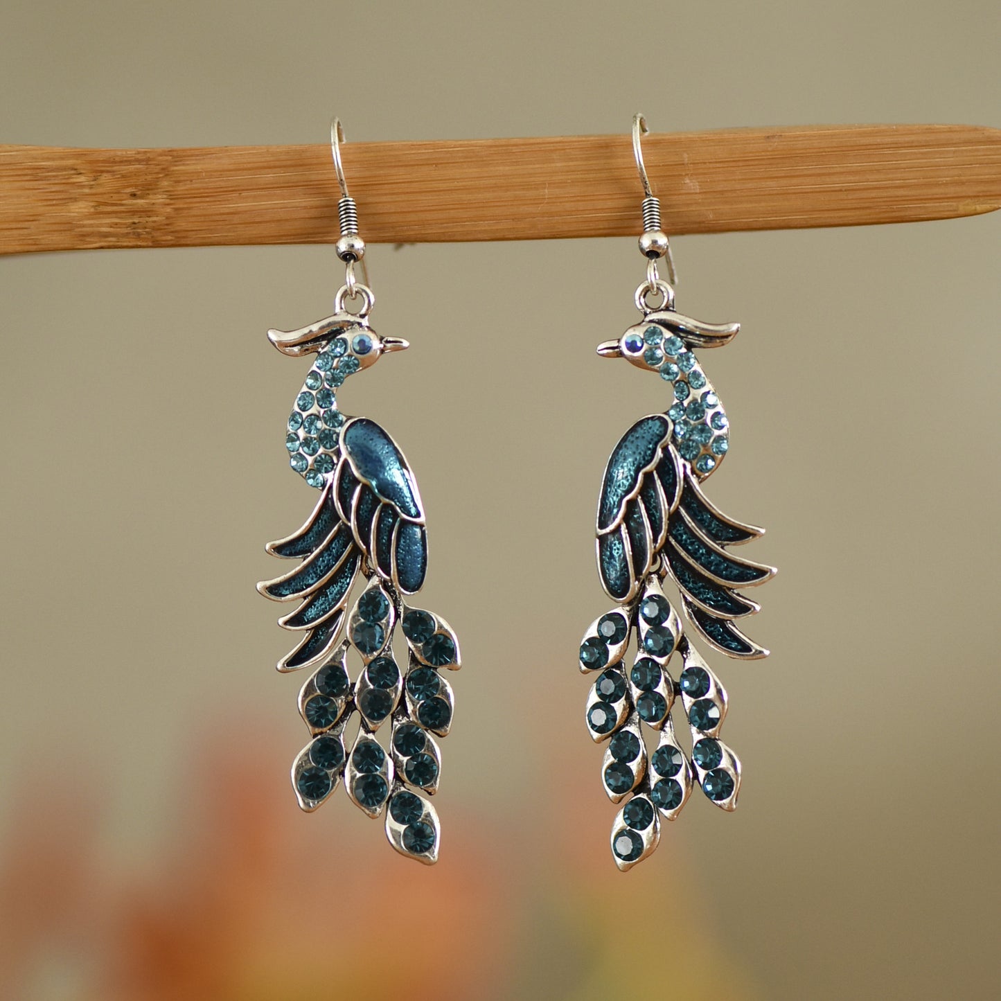 Peacock feather-shaped earrings with blue accents on a wooden stick against a blurred background