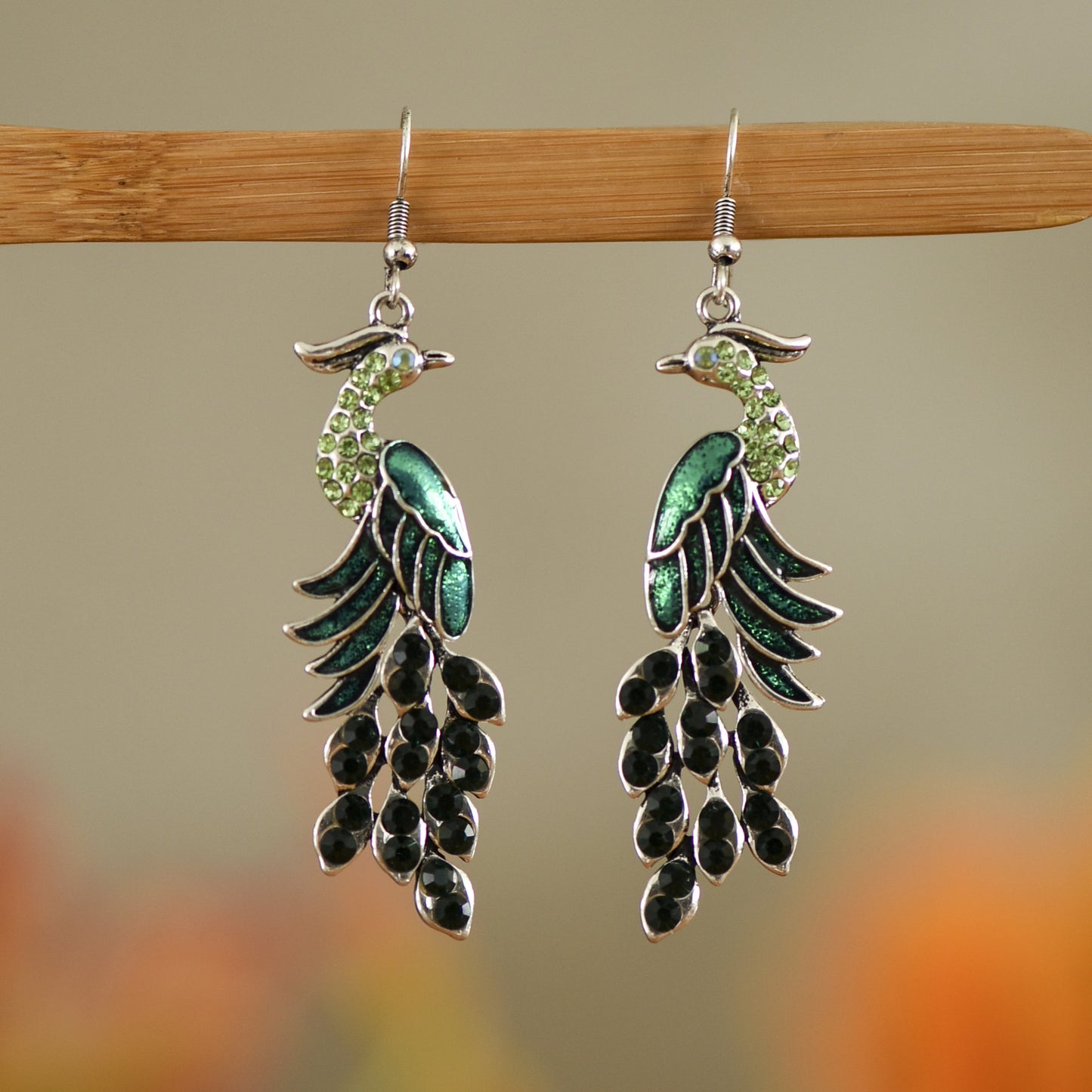 Pair of green and black peacock earrings on a wooden hanger with a blurred nature background.
