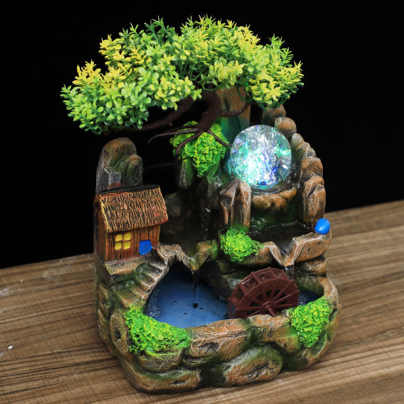 Decorative miniature fountain with a small house, tree, and glowing ball on a wooden surface.