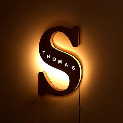Decorative wall light shaped like a letter 'S' with 'THOMAS' text, glowing warmly against a dark background.