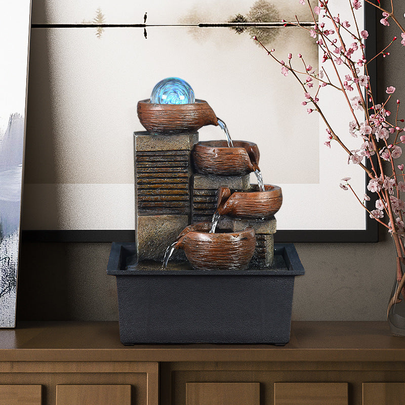 Tabletop water fountain with three bowls on a wooden surface, with a vase of pink flowers in the background.