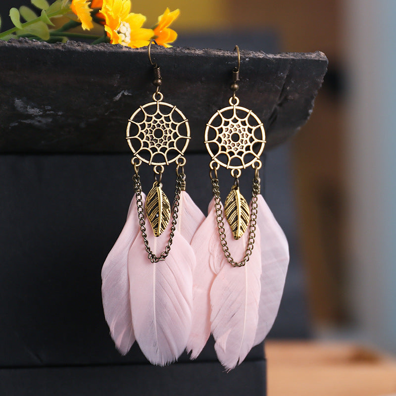 Gold dreamcatcher earrings with pink feathers on a dark surface with yellow flowers in the background.
