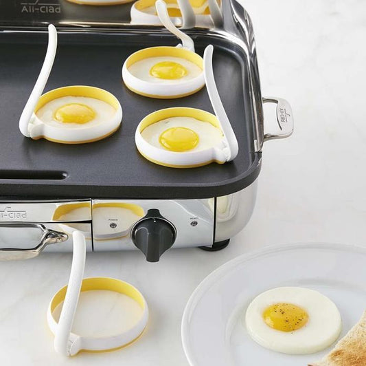 Egg cooking rings on a griddle with eggs being cooked, next to a plate with a cooked egg.