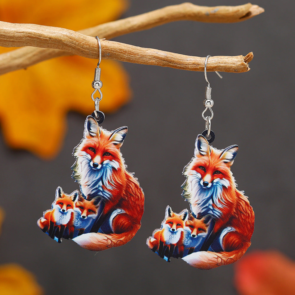 Fox-themed earrings hanging from a branch with a blurred autumn leaf background