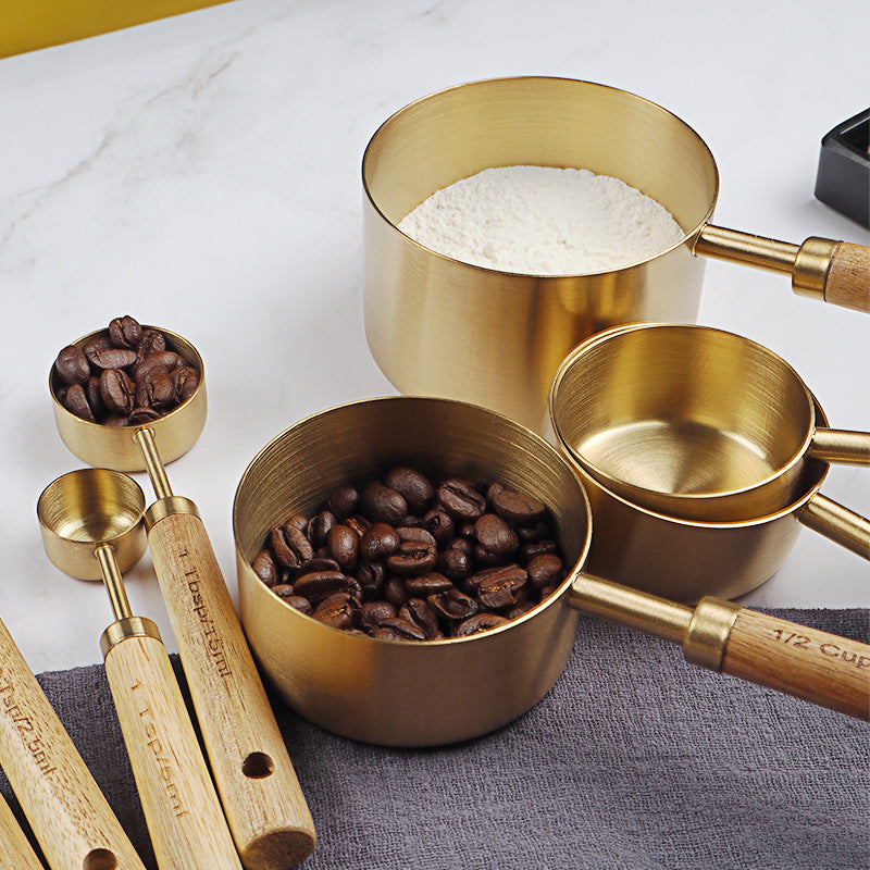 Set of gold measuring cups with wooden handles containing coffee beans and flour on a white surface.