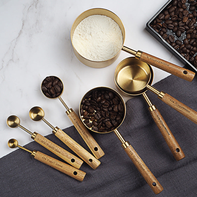 Set of brass and wooden measuring spoons with coffee beans, flour, and a tray of coffee beans on a marble surface.