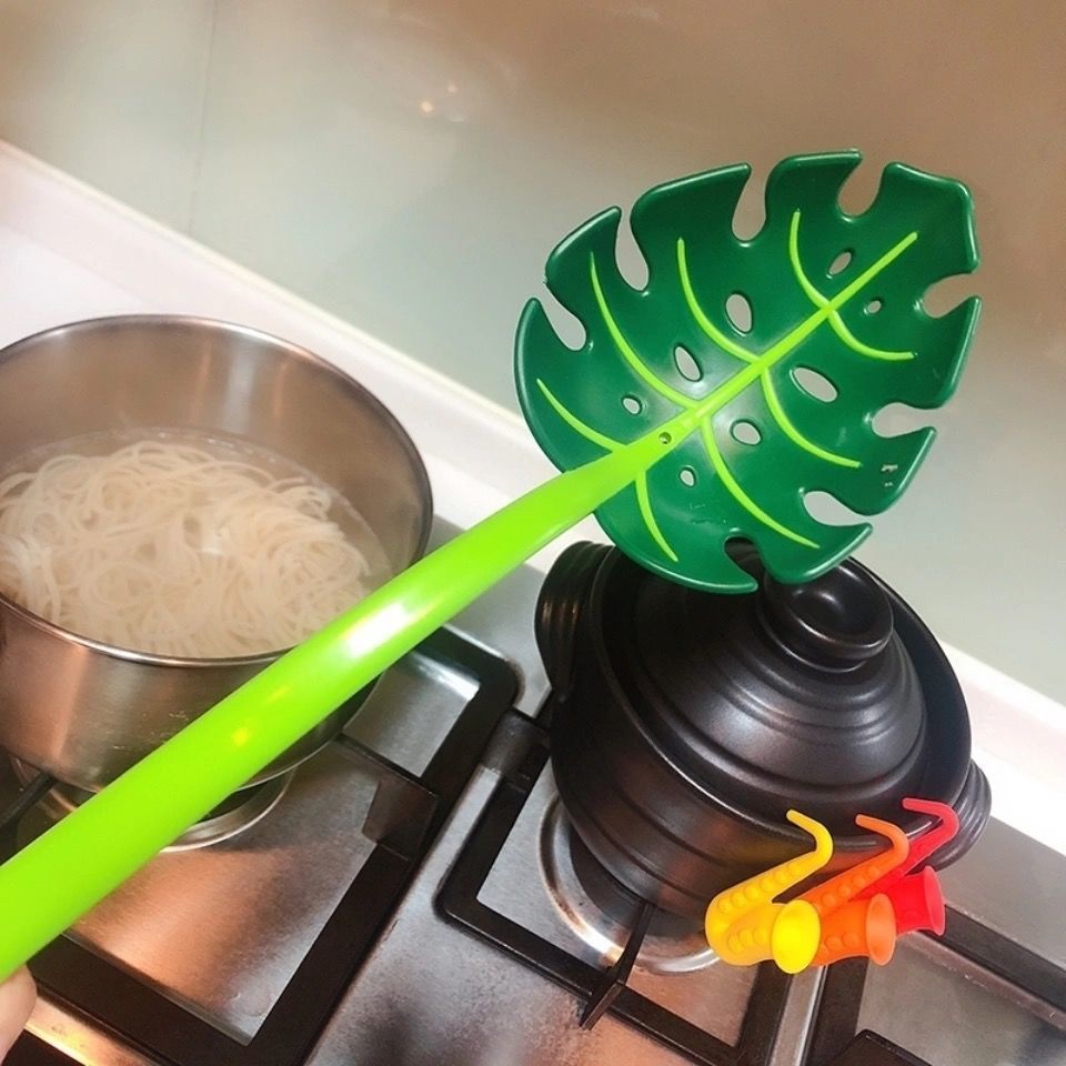 Green leaf-shaped ladle over a pot of boiling noodles on a stove.