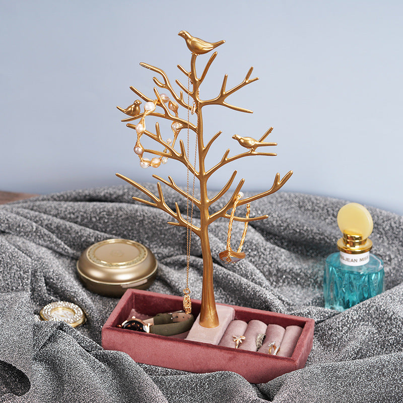 Gold tree-shaped jewelry stand with birds on top, holding necklaces, on a gray fabric surface.