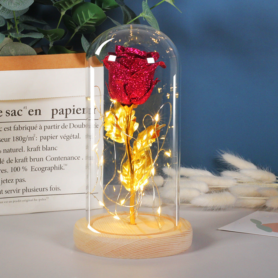 Red rose in a glass dome with LED lights on a wooden base, placed on a surface with a blurred background.