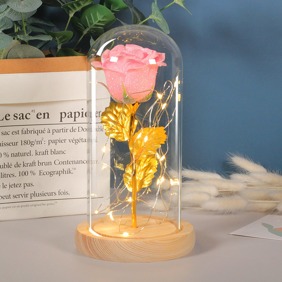 Decorative setup with a pink rose and gold leaf inside a glass dome on a wooden base, against a blurred background.