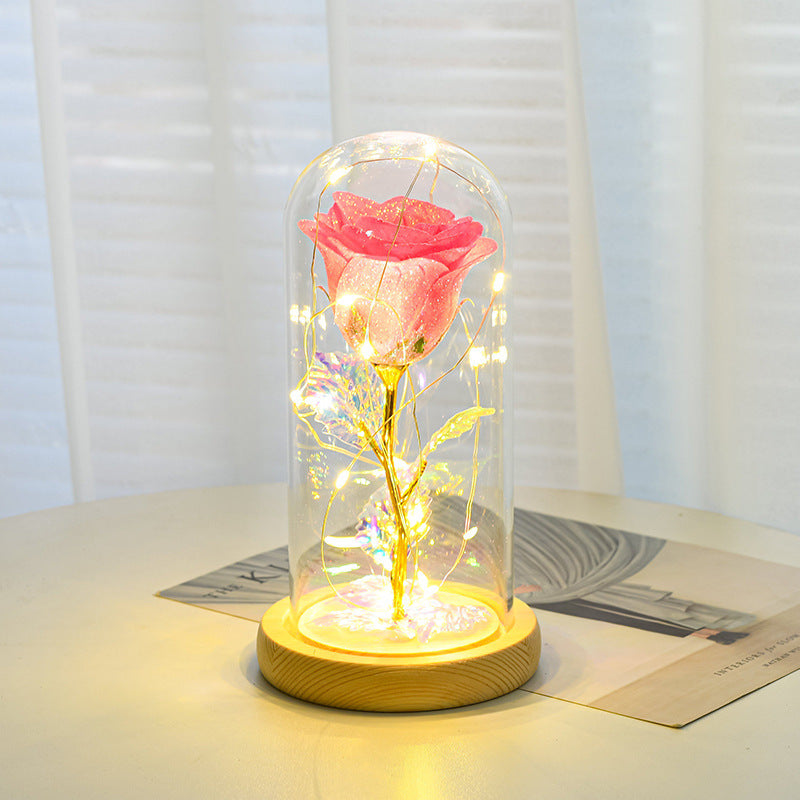 Pink rose in a glass dome with LED lights on a wooden base, placed on a surface with a light background.