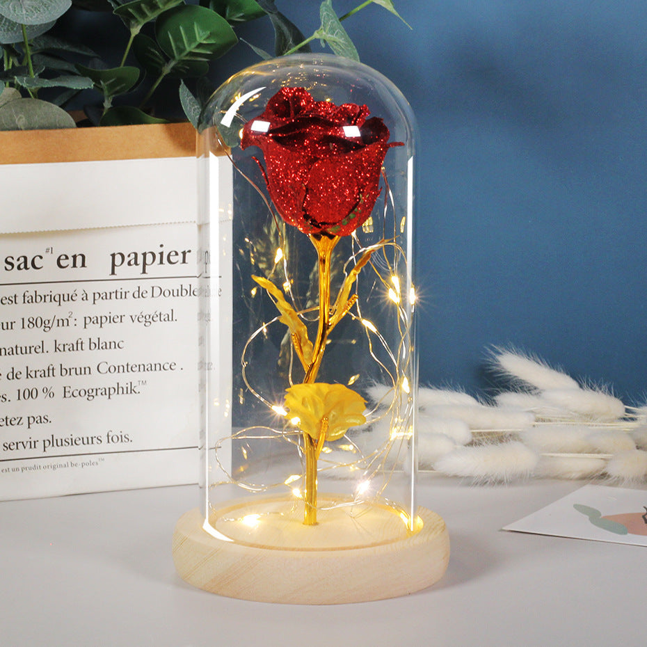 Red and gold rose in a glass dome with LED lights on a wooden base, against a blue wall with plants and feathers.