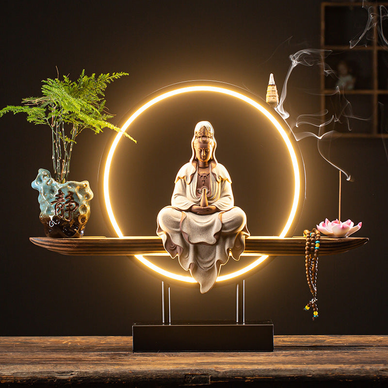 Decorative setup with a statue of a meditating figure, illuminated circle, plants, and incense on a wooden surface.