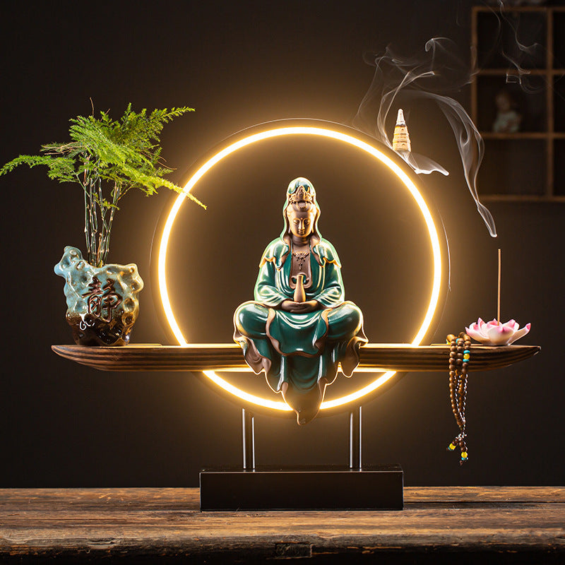 Decorative setup with a statue of a seated figure, plants, and a glowing circular light on a wooden surface.