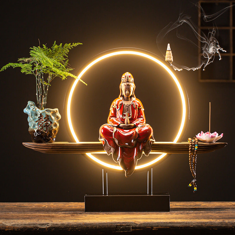 Decorative setup with a red Buddha statue, plants, and incense on a wooden surface.