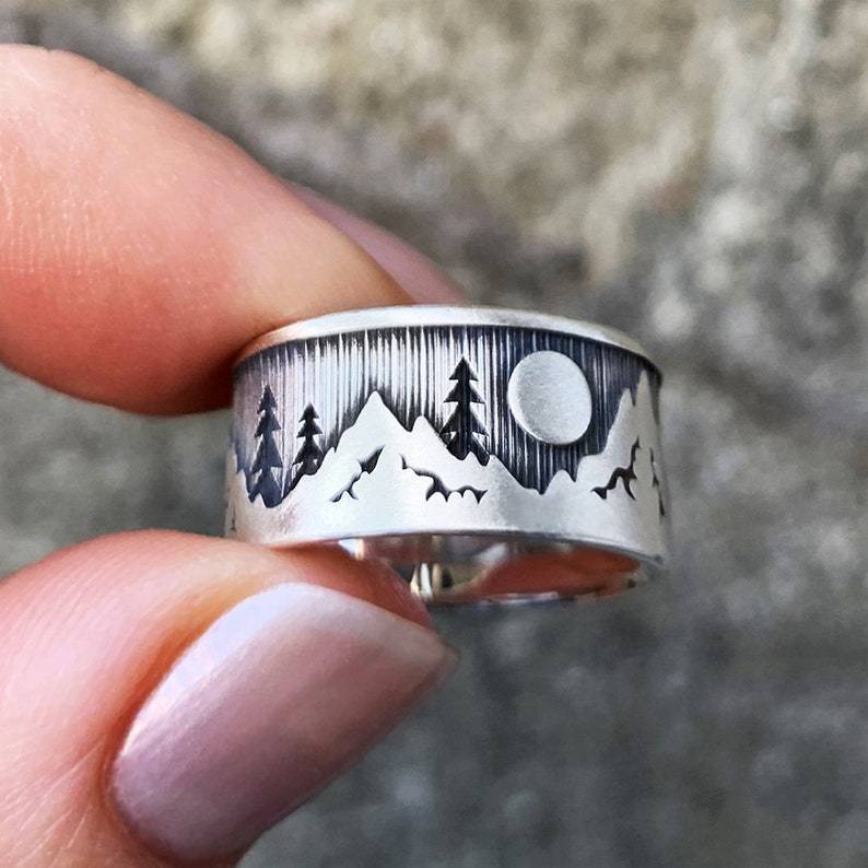 Silver ring with nature-themed design held between fingers against a blurred background