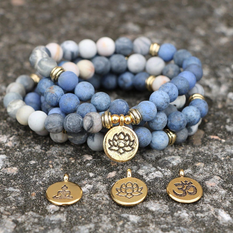Set of blue and gray beaded bracelets with gold lotus and om charms on a stone surface.