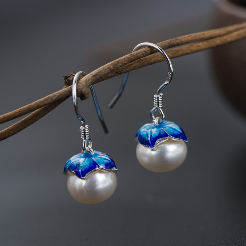 Pearl earrings with blue floral designs hanging on a branch against a dark background
