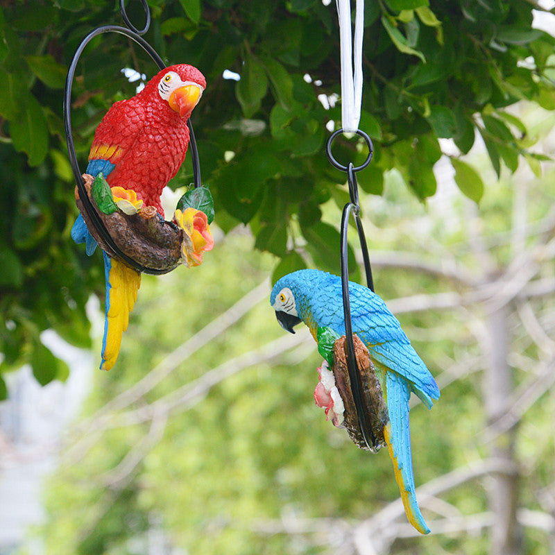 Two colorful parrot figurines hanging from a branch with a blurred green foliage background.