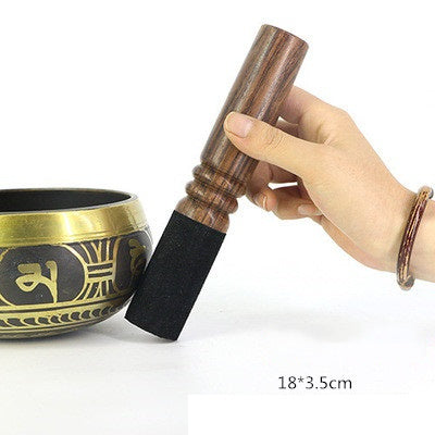 Hand holding a wooden singing bowl striker next to a singing bowl on a white background