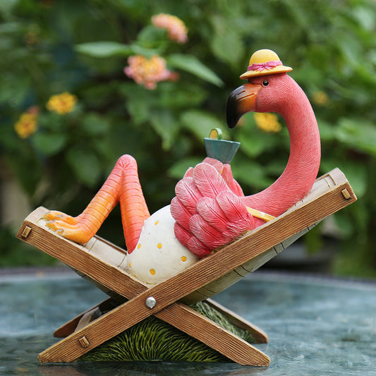 Pink flamingo figurine with a hat and drink on a lounge chair against a blurred green background