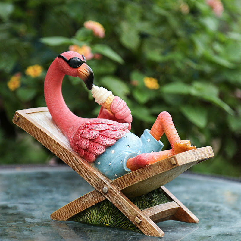 Pink flamingo figurine lounging on a deck chair with sunglasses and a drink, against a blurred green foliage background.