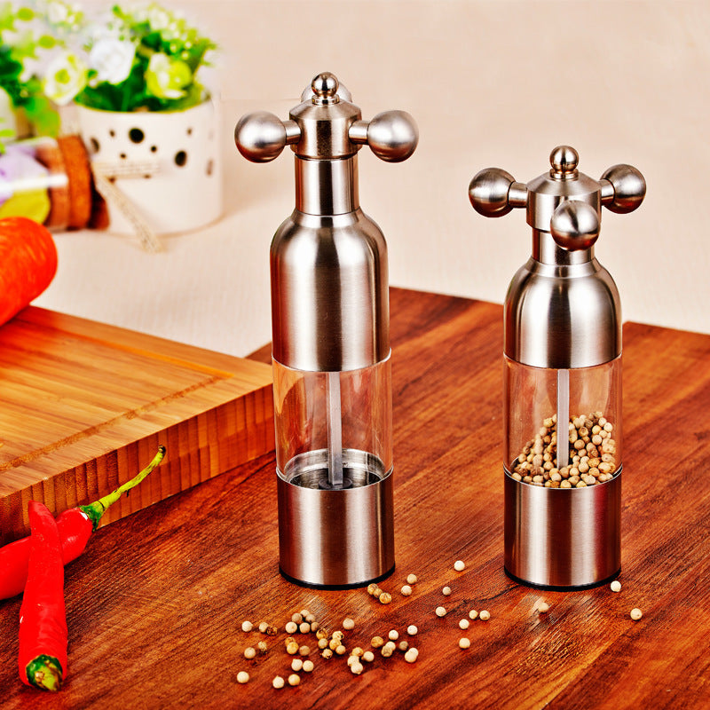 Two silver pepper mills on a wooden surface with spices around.